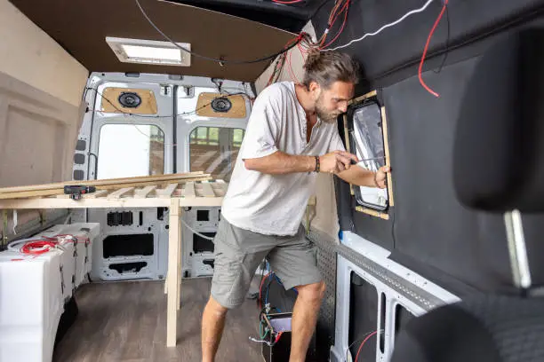 Interior view. A man is converting a van into a caravan. He is tightening a screw in the side window.