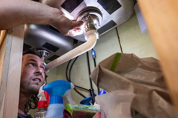 A man uses a flashlight to inspect or repair plumbing beneath a kitchen sink, surrounded by household cleaning supplies. This candid, real-life scene highlights home maintenance, DIY repairs, and responsible homeownership.
